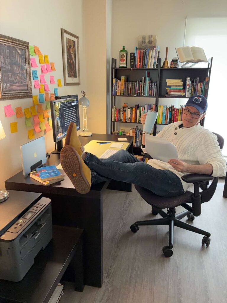 Corey wears an Oilers cap and reads a paper manuscript with his feet upon a desk arrayed with four books by Canadian authors, a lamp, and his computer. Behind his desk are two bookshelves filled with books and a print of Cot's Springtime hangs upon the wall.